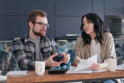 Casal analisa documentos e conversa sobre finanças sentado à mesa da cozinha.
