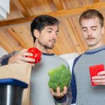 Dois homens organizam compras de supermercado em uma cozinha enquanto consultam um celular.