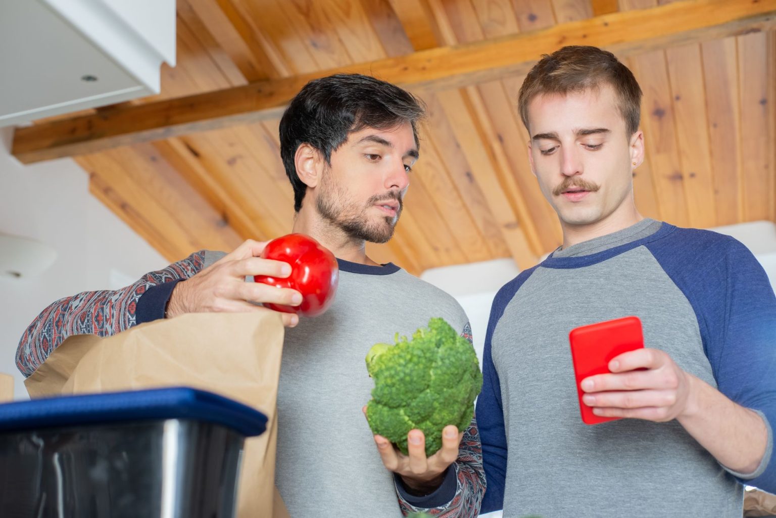 Dois homens organizam compras de supermercado em uma cozinha enquanto consultam um celular.