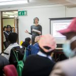Grupo diverso participa de uma reunião comunitária enquanto uma pessoa apresenta informações diante de uma tela.