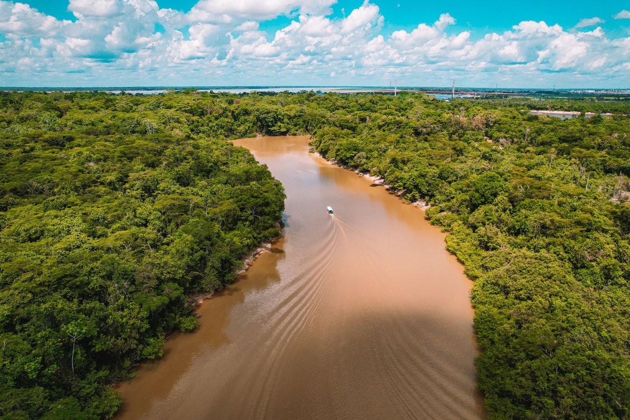 Vista aérea de uma embarcação navegando por um rio barrento cercado pela floresta amazônica.